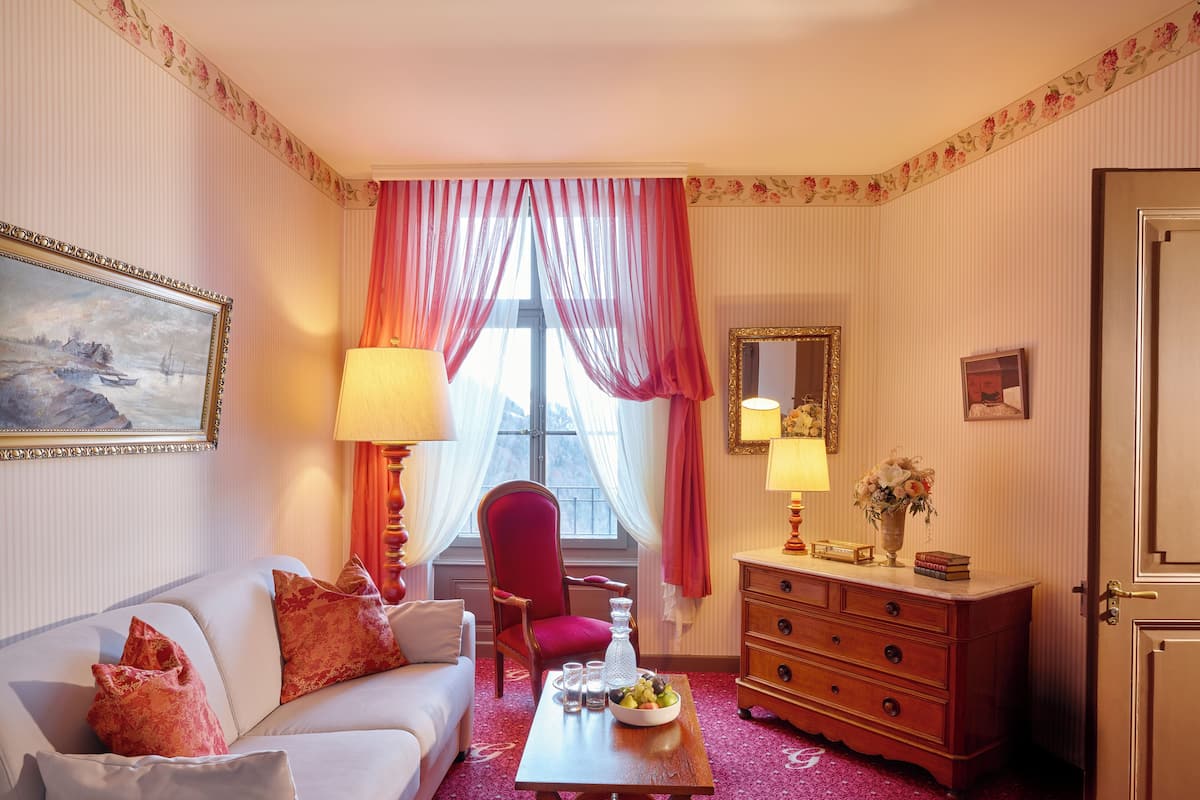 Quarto de hotel com sofá branco, cadeira vermelha, cômoda de madeira e mesa com uma fruteira.