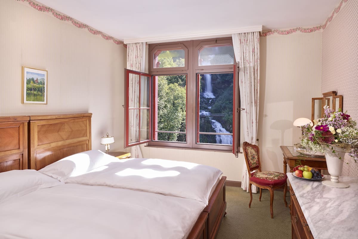Um quarto com uma cama grande, uma mesinha de cabeceira de madeira, uma escrivaninha com espelho, uma cadeira e uma janela com vista para uma cascata.