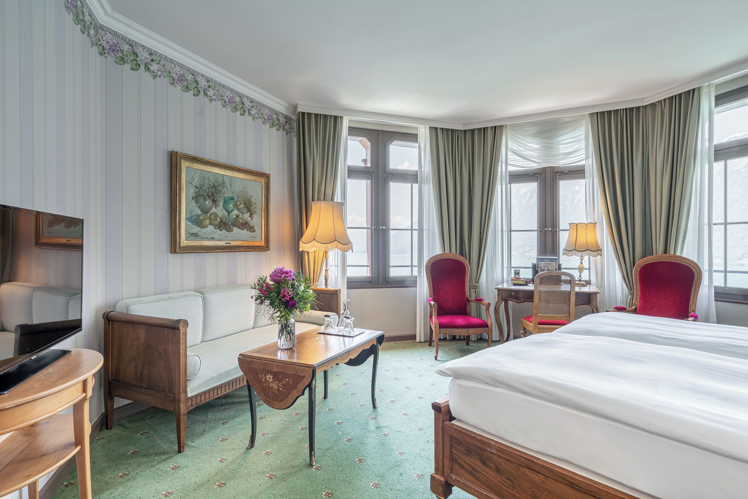 A hotel room with a bed, a desk, a chair, a sofa, a TV, and a painting on the wall.