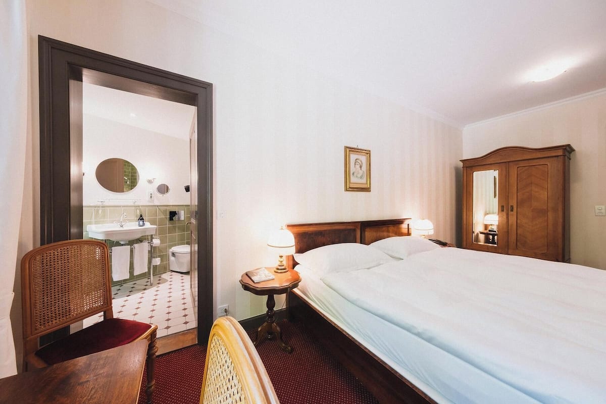 Quarto de hotel com cama, mesa de cabeceira com abajur, guarda-roupa de madeira, uma cadeira, banheiro com pia e espelho, e piso ladrilhado.