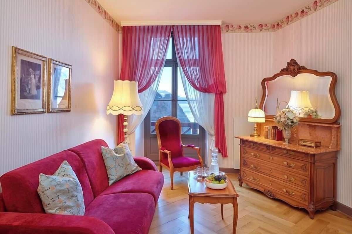 Quarto com sofá vermelho, cómoda de madeira, cadeira e mesa com uma tigela de fruta.