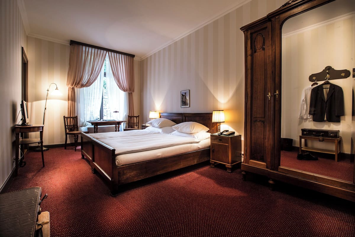 Quarto de hotel com uma cama grande, um guarda-roupa, uma secretária com abajur e um espelho.