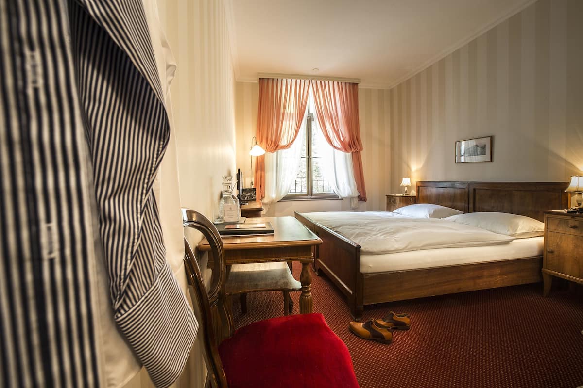 Quarto de hotel com cama, escrivaninha, cadeira, janela com cortinas e um quadro na parede.