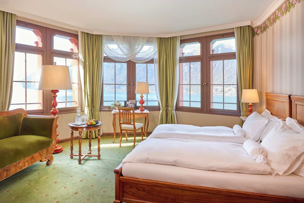 Quarto de hotel com cama, poltrona, mesa e vista para um lago através de grandes janelas.