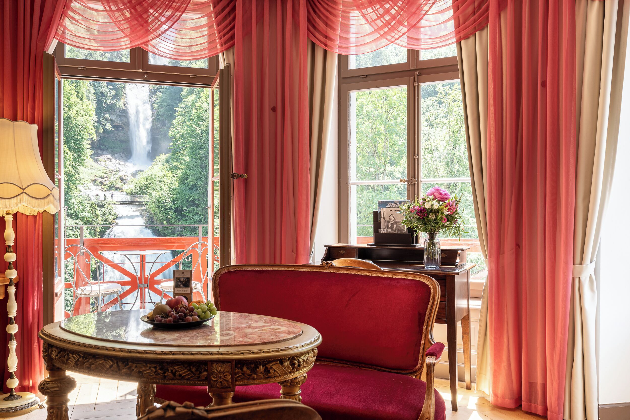 A room with a view of a waterfall, a round table with fruit, a red chair, and a piano.