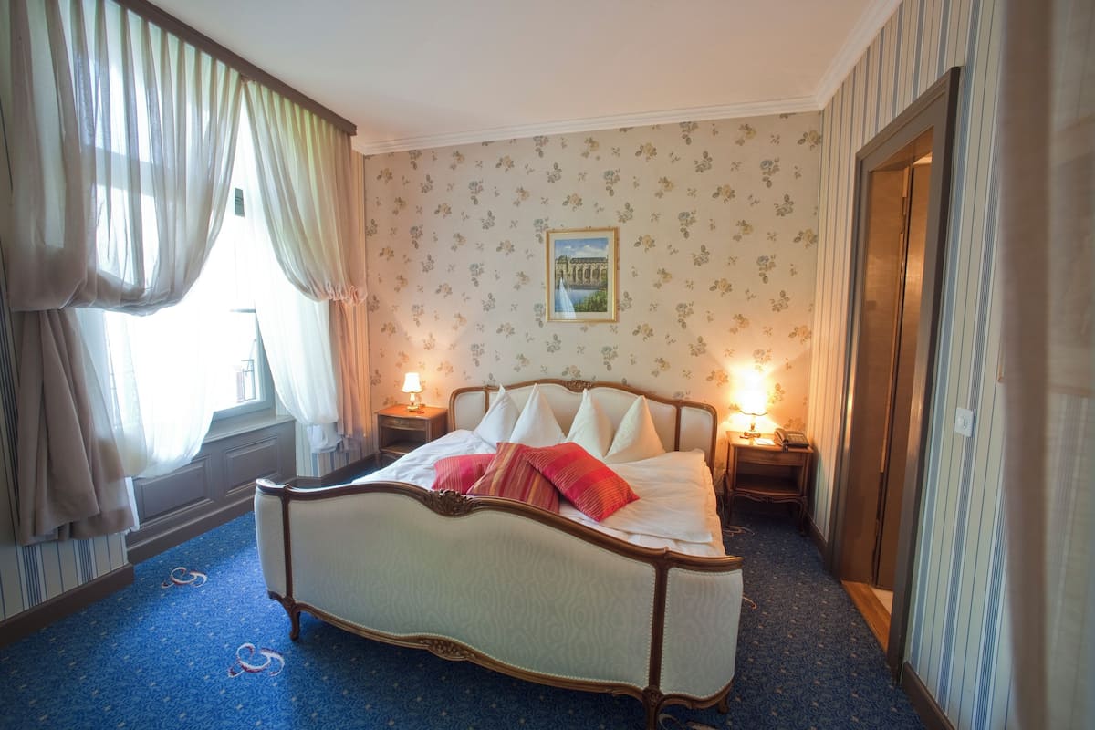 Um quarto com uma cama grande, duas mesas de cabeceira com luminárias, uma janela com cortinas e uma parede com papel de parede floral.