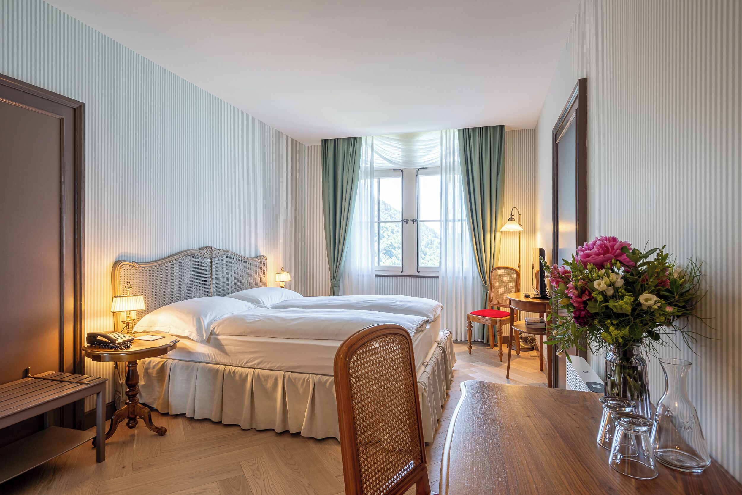 Ein Hotelzimmer mit Doppelbett, Schreibtisch mit Stuhl, Nachttisch mit Lampe und einem großen Fenster mit Vorhängen.