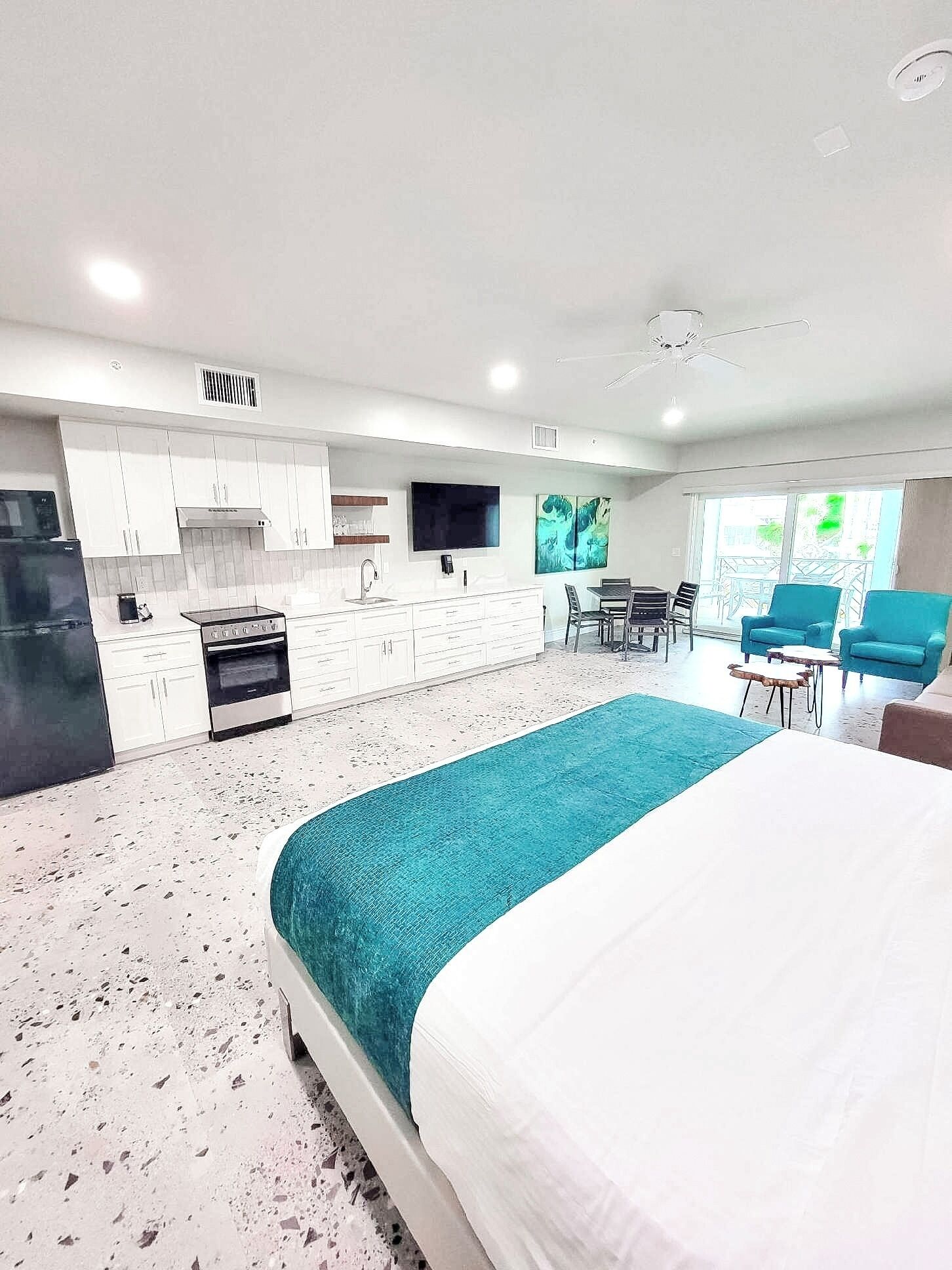 A modern hotel room with a large bed, a kitchenette, a dining area, and a view of the outdoors.