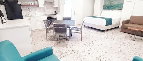 A compact living space with a kitchenette, a dining area, a bed, and two teal armchairs.