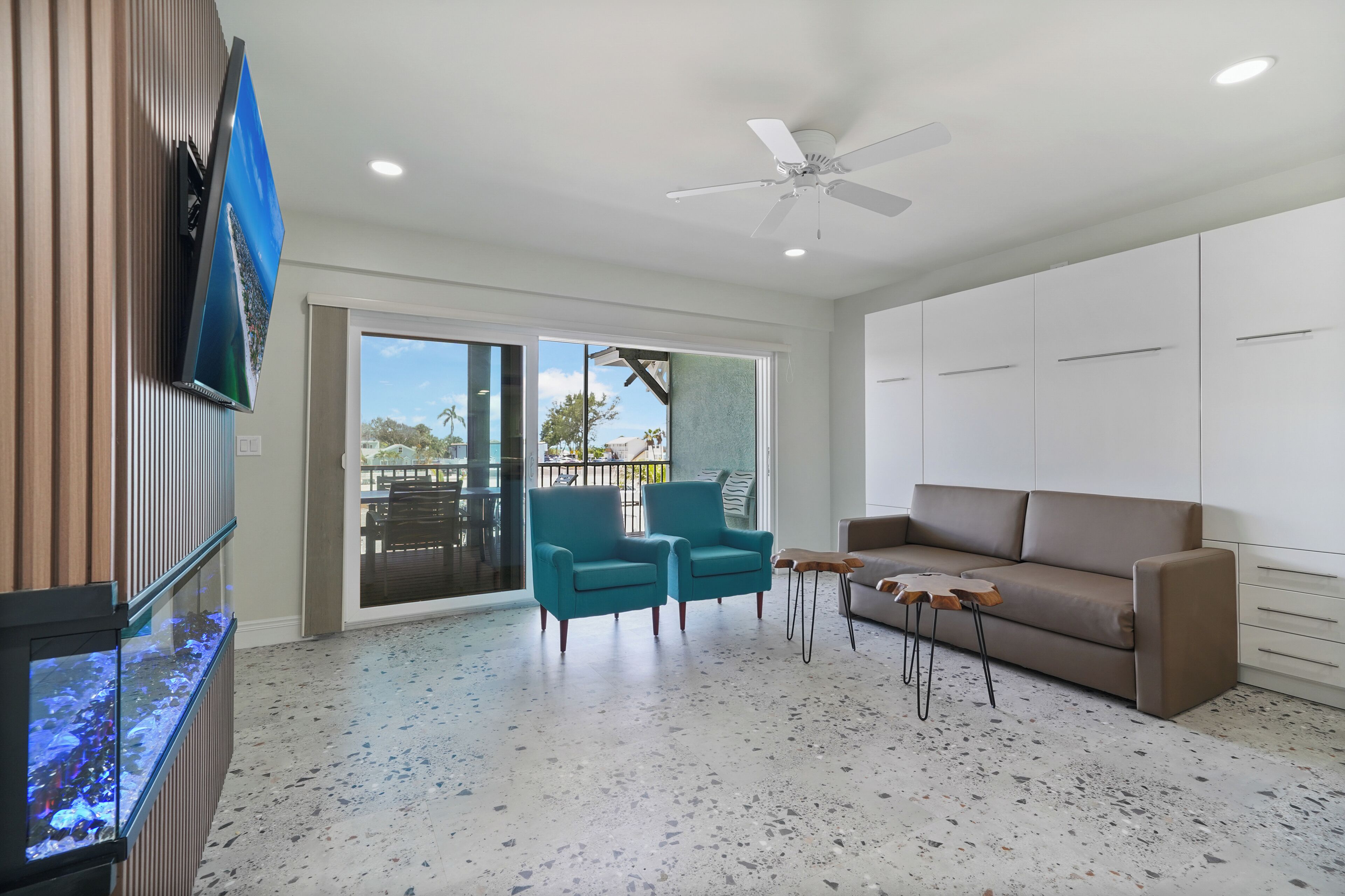 A spacious living area with a large TV, teal armchairs, a brown sofa, and a ceiling fan.