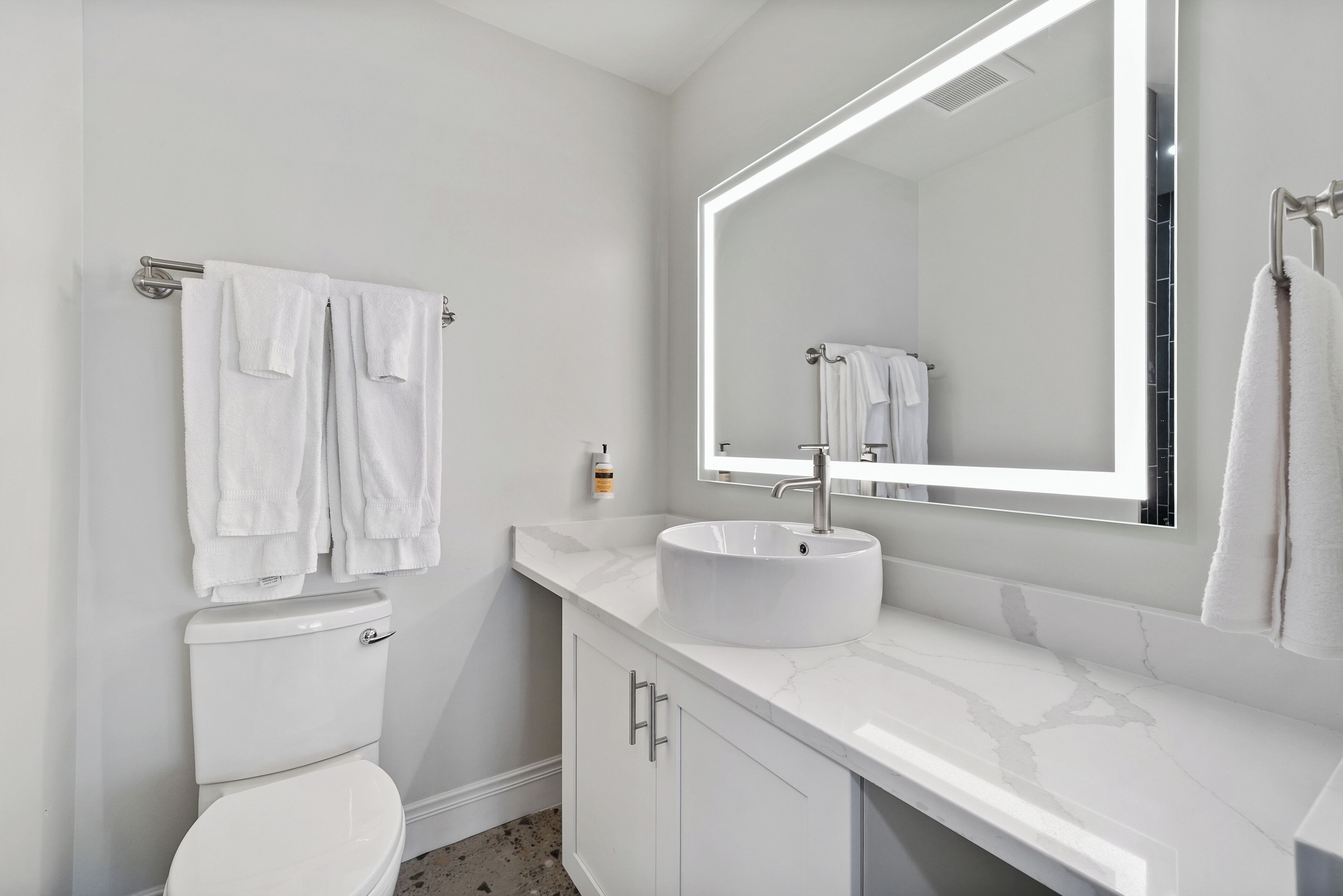 Une salle de bain moderne avec des toilettes blanches, un lavabo doté d’un plan de travail en marbre et un grand miroir avec éclairage intégré.