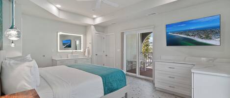 A modern bedroom with a bed, a ceiling fan, a large TV, and a balcony with a view of the ocean.