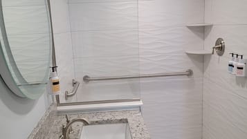 Un baño moderno con lavamanos de encimera de granito, cabina de ducha con mampara de vidrio y zona de ducha con azulejos blancos.