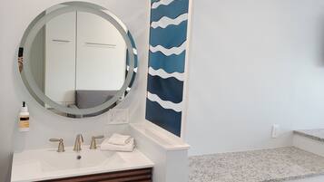 A bathroom with a white vanity, a round mirror, a wall-mounted sink, a towel, and a blue upholstered bench.