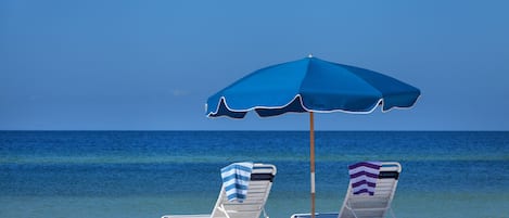 Two beach chairs under a blue umbrella on a sandy beach with the ocean in the background.