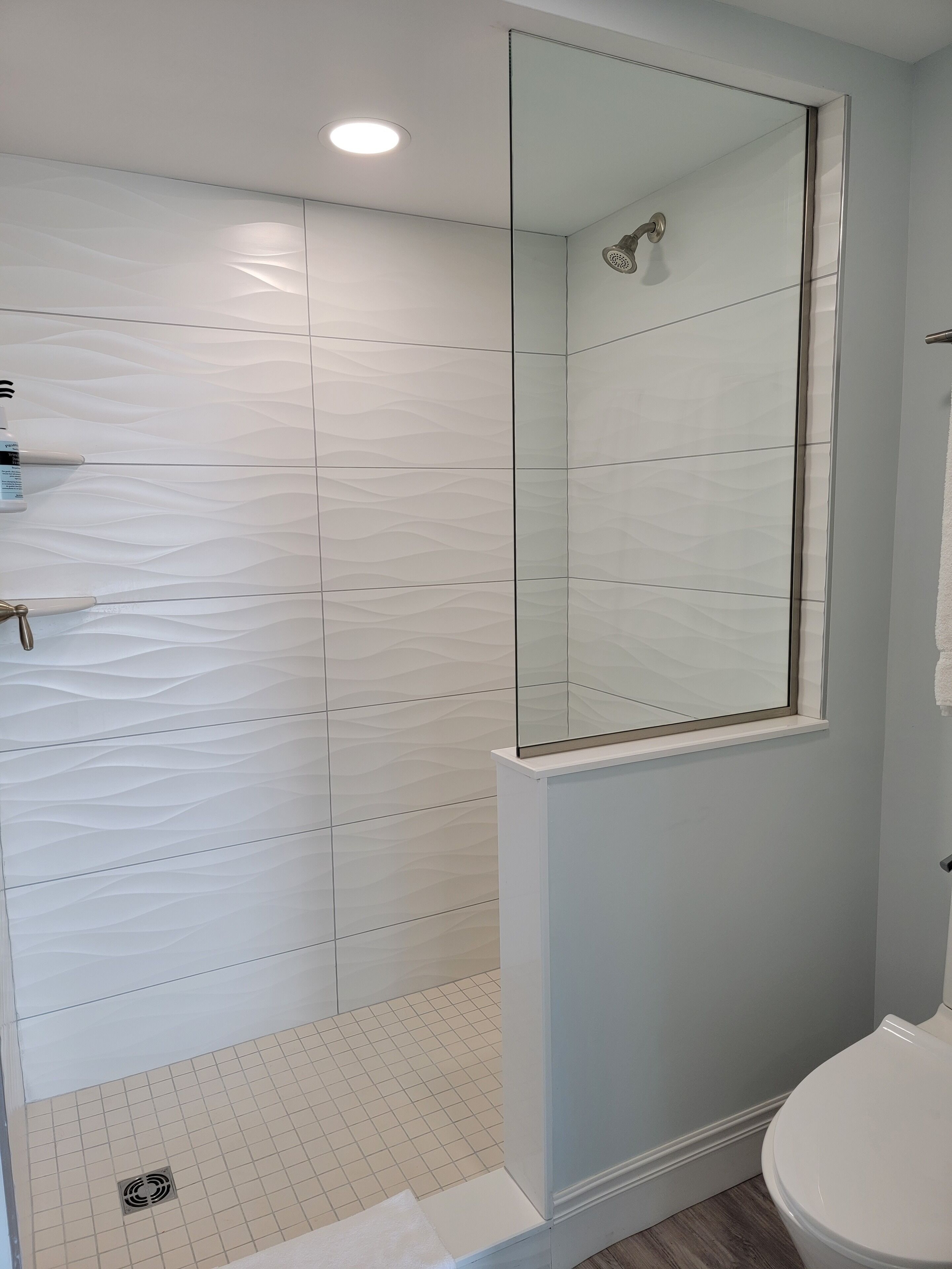 Un baño con cabina de ducha de vidrio, paredes con azulejos blancos, inodoro blanco y un estante pequeño con artículos de aseo.