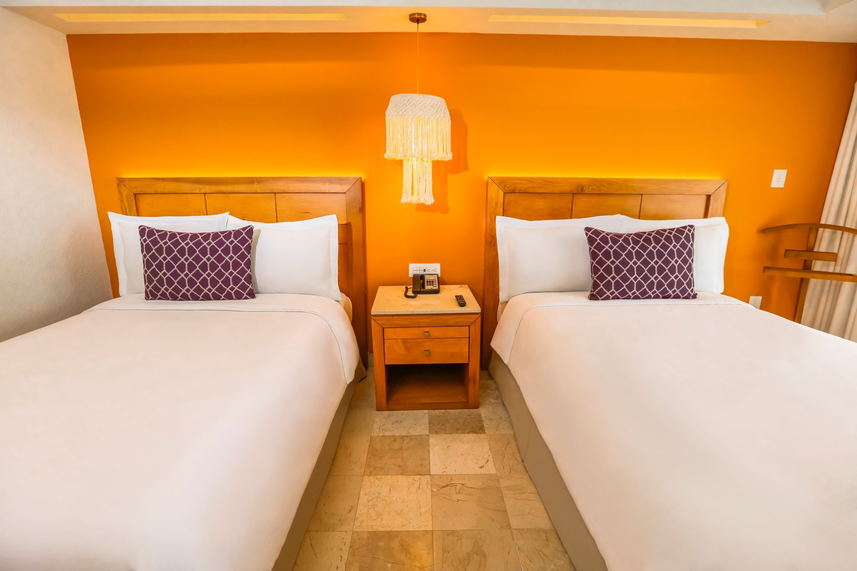 A hotel room with two beds, each with white linens and patterned pillows. There is a nightstand with a lamp between the beds.