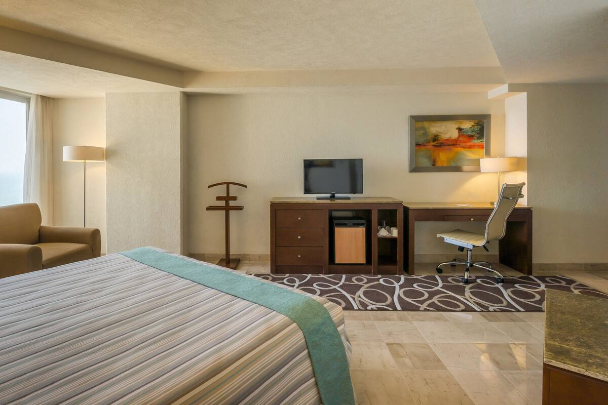 A hotel room with a bed, a desk, a chair, a television, and a painting on the wall.