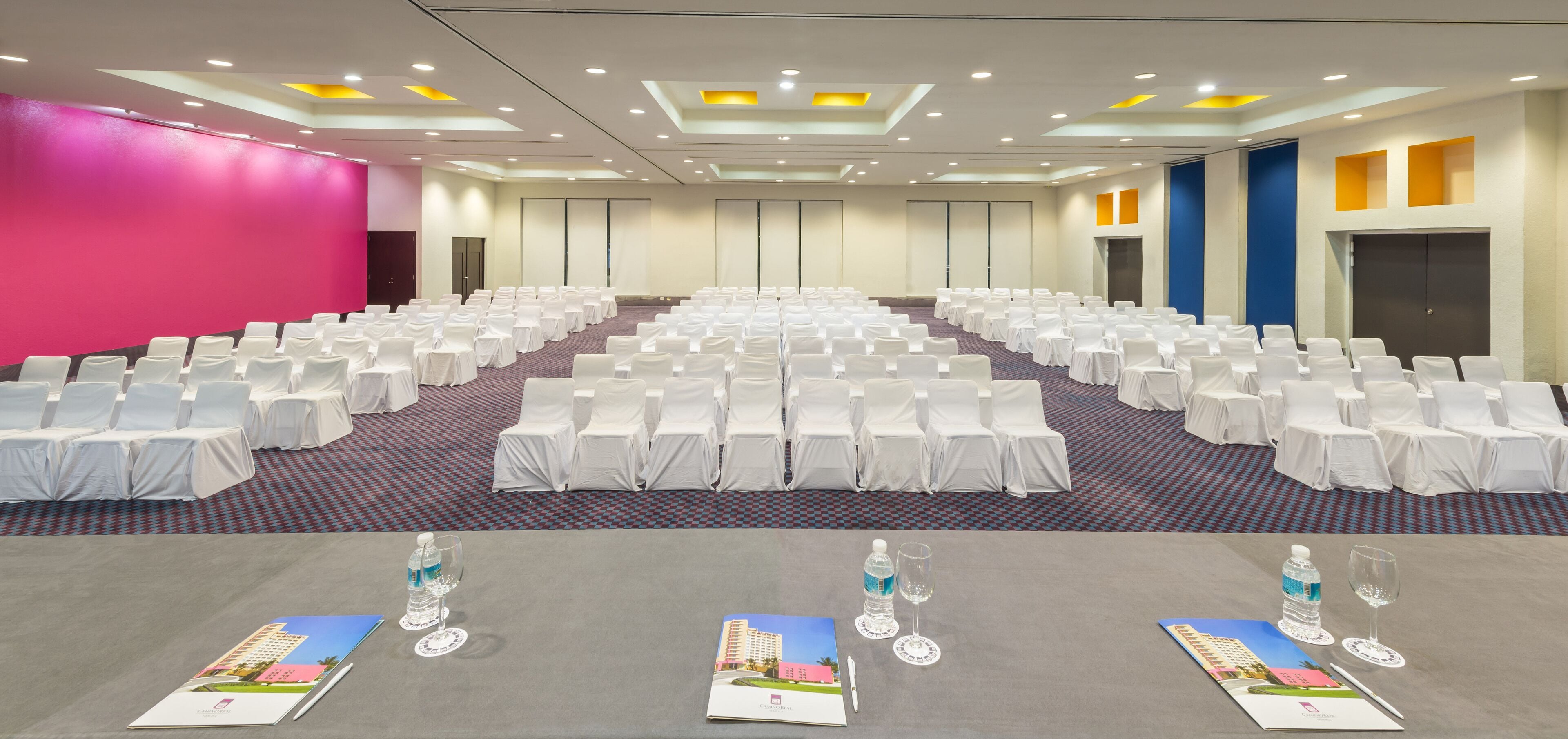 Une salle de conférence avec des rangées de chaises blanches, des tables avec des bouteilles d’eau et des verres, et des brochures d’information.