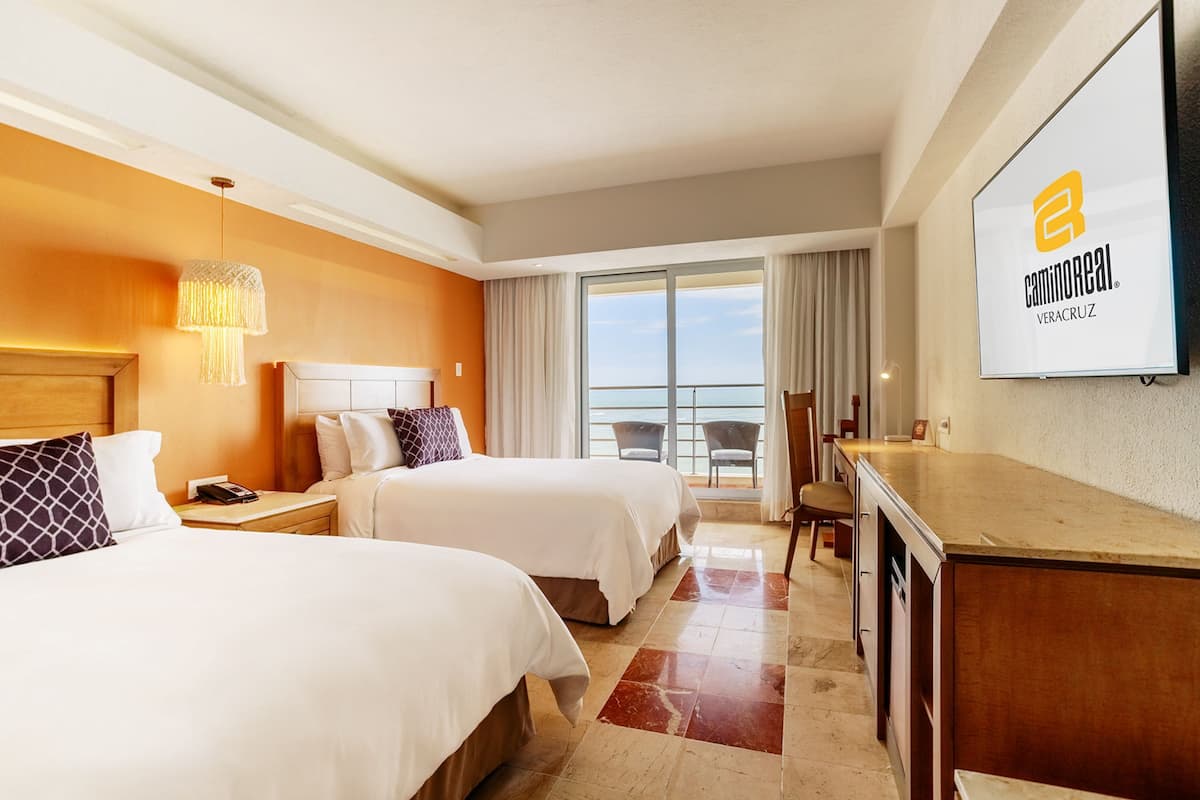 A hotel room with two beds, a desk, a chair, a TV, and a balcony with a view.