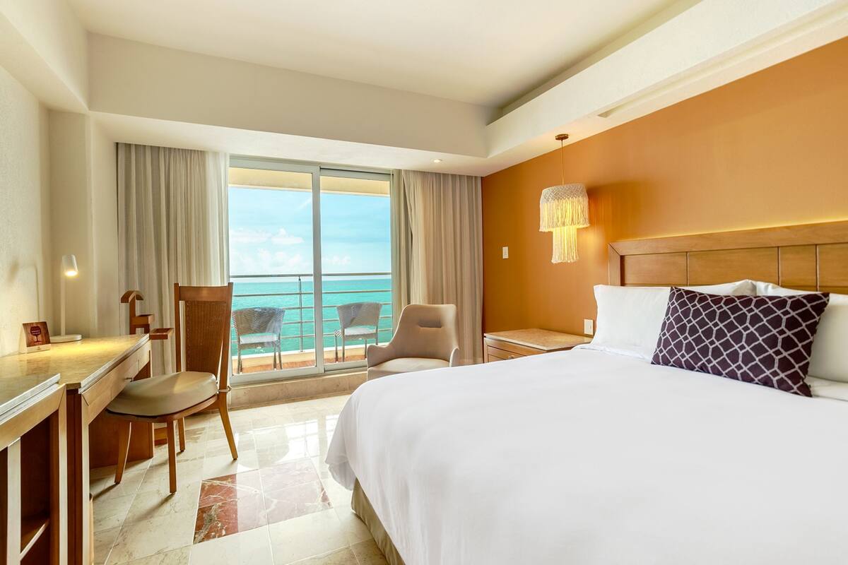 A hotel room with a large bed, a desk, a chair, and a view of the ocean through a sliding door.
