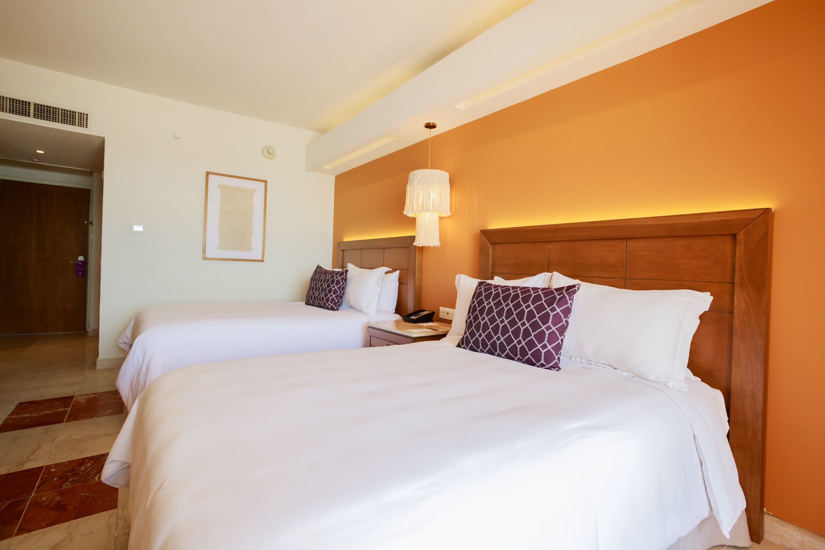 A hotel room with two beds, a wooden headboard, a nightstand, a framed picture on the wall, and a hanging light fixture.