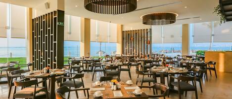 A spacious dining area with multiple tables and chairs, large windows offering a view of the sea, and modern pendant lighting fixtures.