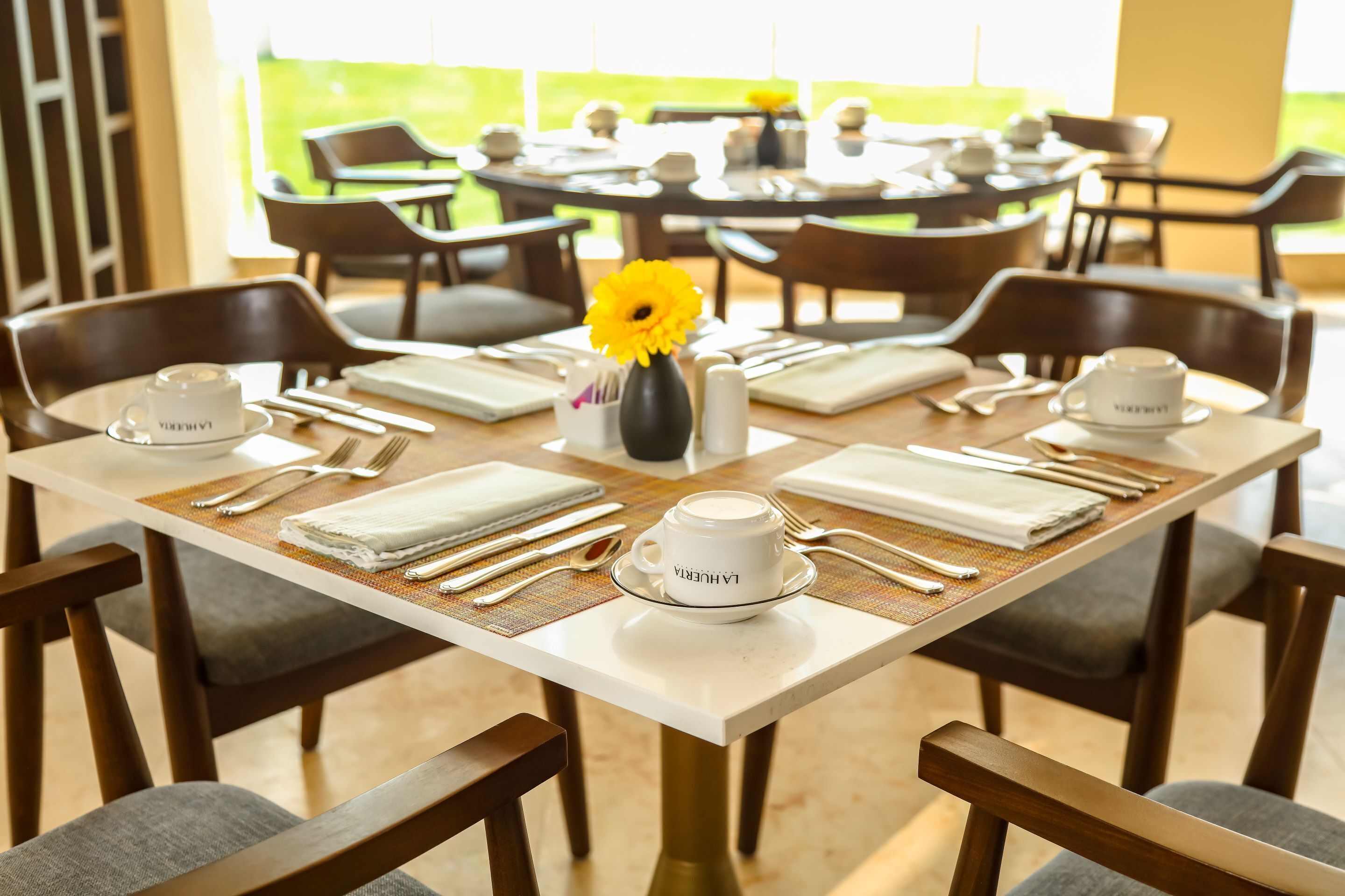Un espace repas avec des tables dressées pour quatre personnes, chacune agrémentée d’une nappe blanche, de couverts et d’un petit vase contenant une fleur jaune.