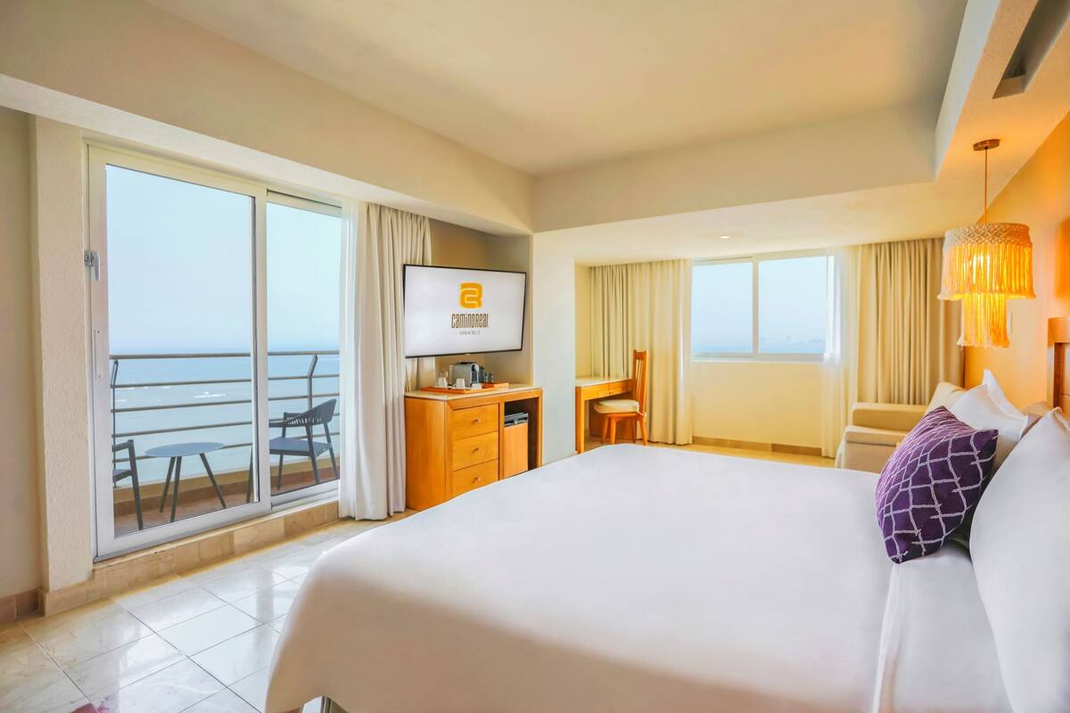 A hotel room with a large bed, a desk with a computer, a chair, and a view of the sea through a sliding door.
