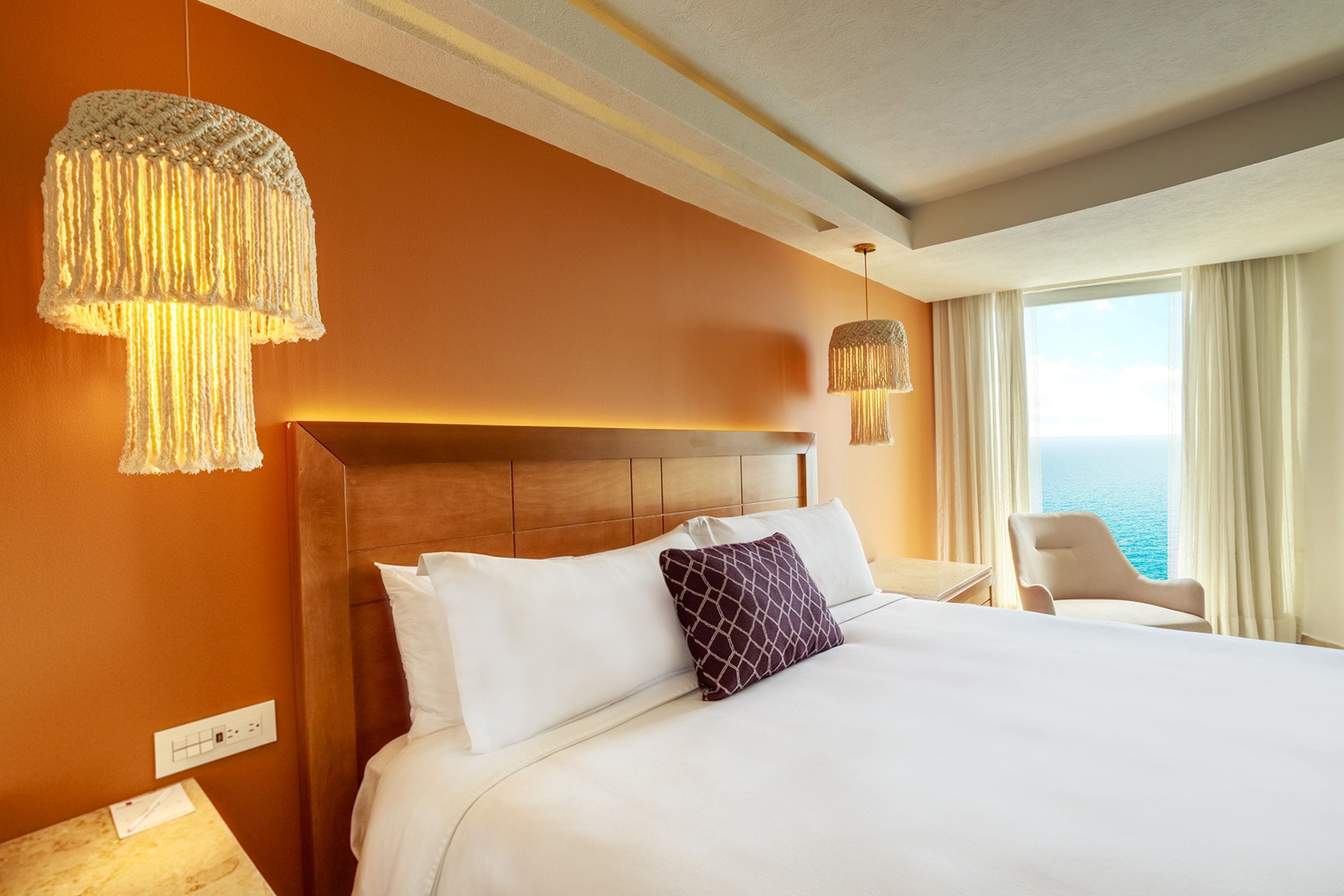 Quarto de hotel com uma cama grande, cabeceira de madeira, vista para o mar pela janela e luminária suspensa.