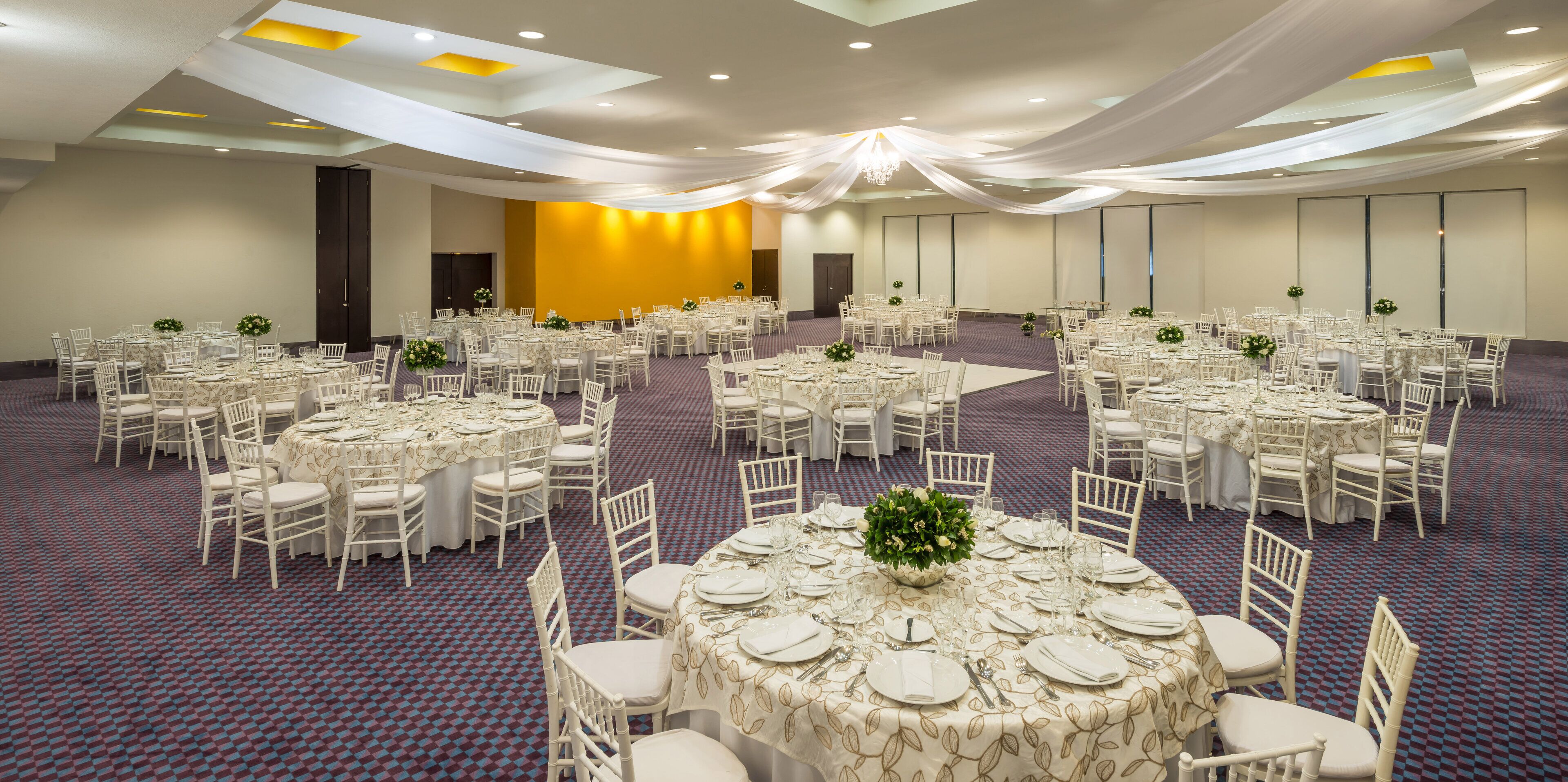 Un amplio salón de banquetes con mesas redondas, sillas blancas y centros de mesa con flores.