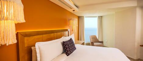 Habitación de hotel con una cama grande, cabecera de madera, vista al mar y una zona de estar con mesa y sillas.