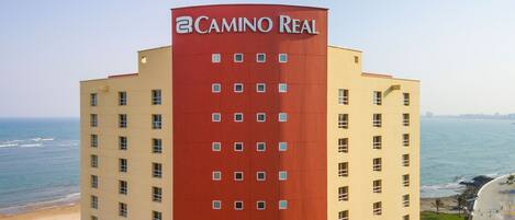 A multi-story building with a red and beige facade, named "Camino Real," featuring a coastal view with the ocean and beach in the background.