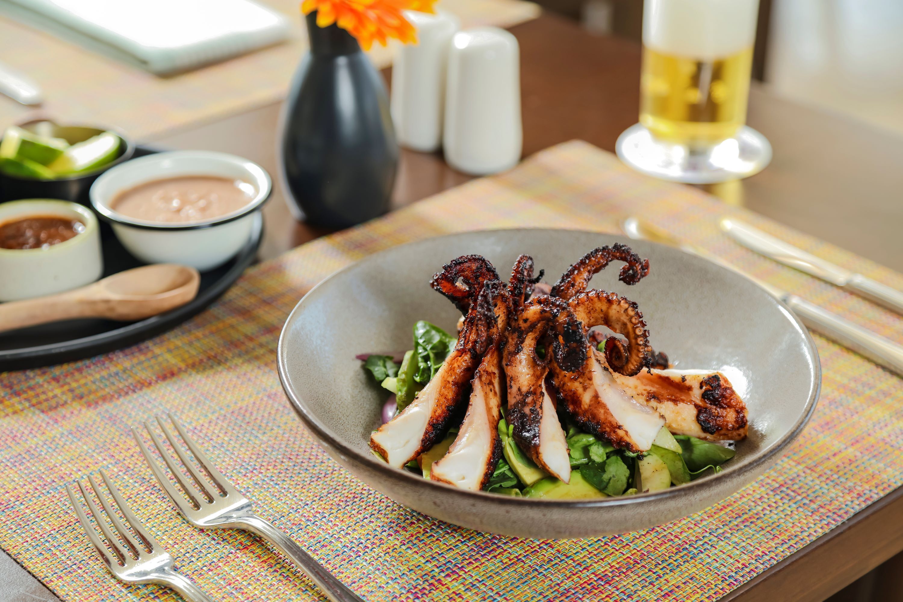 Une assiette de poulpe grillé accompagnée d’une salade, posée sur une table avec une fourchette et un couteau, un vase de fleurs et un verre de vin.