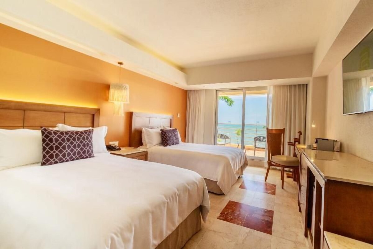 A hotel room with two beds, a desk, a chair, and a balcony with a view of the ocean.