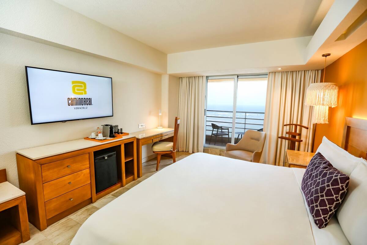 A hotel room with a bed, desk, chair, and a TV displaying the hotel’s logo.