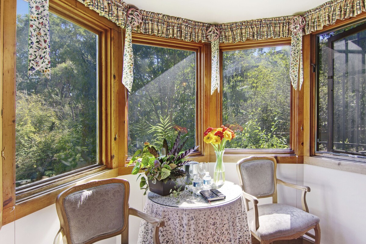 A corner room with a round table, two chairs, a vase with flowers, and a view of greenery through large windows.