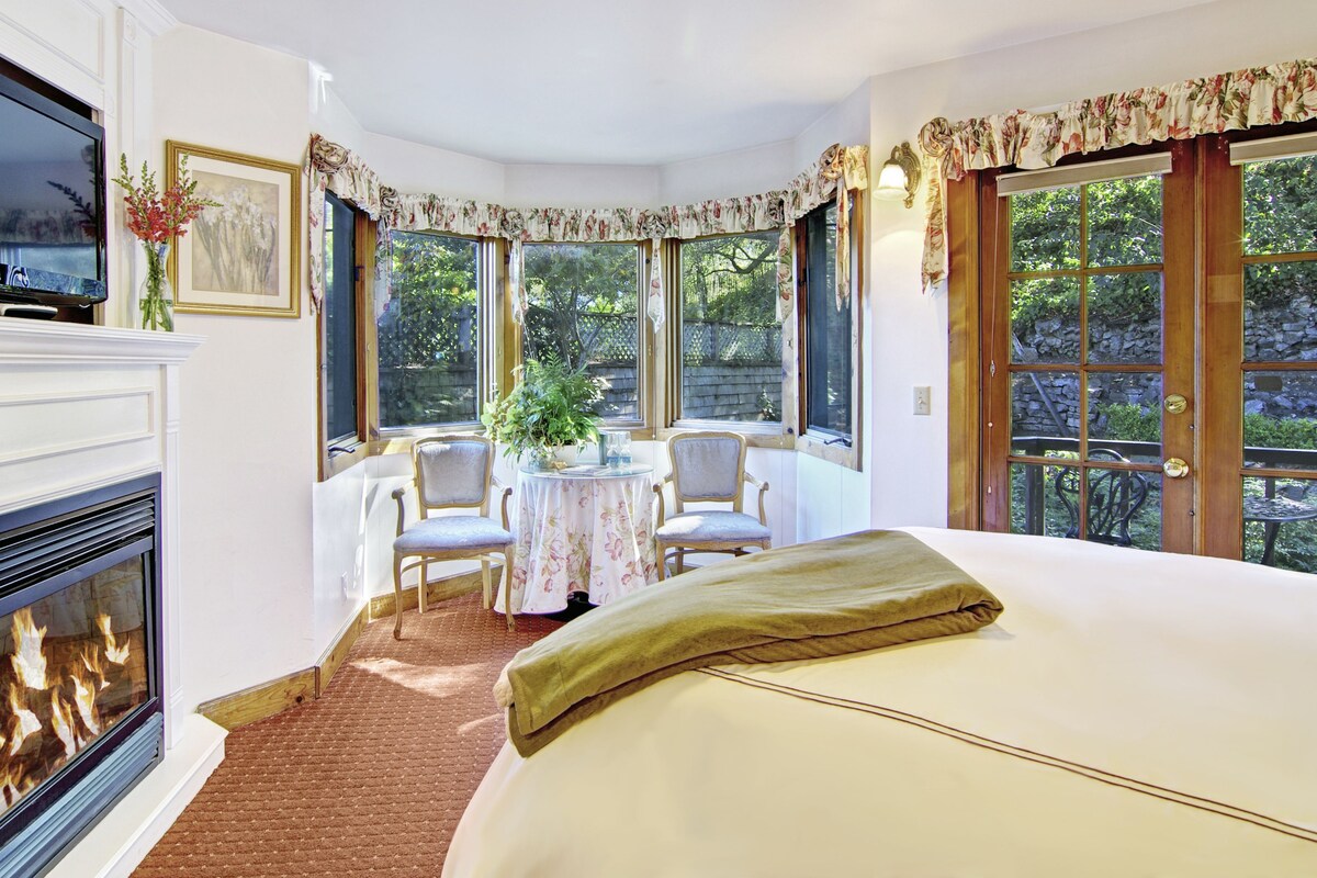 A hotel room with a fireplace, a television, a dining table with chairs, and large windows offering a view of the outdoors.
