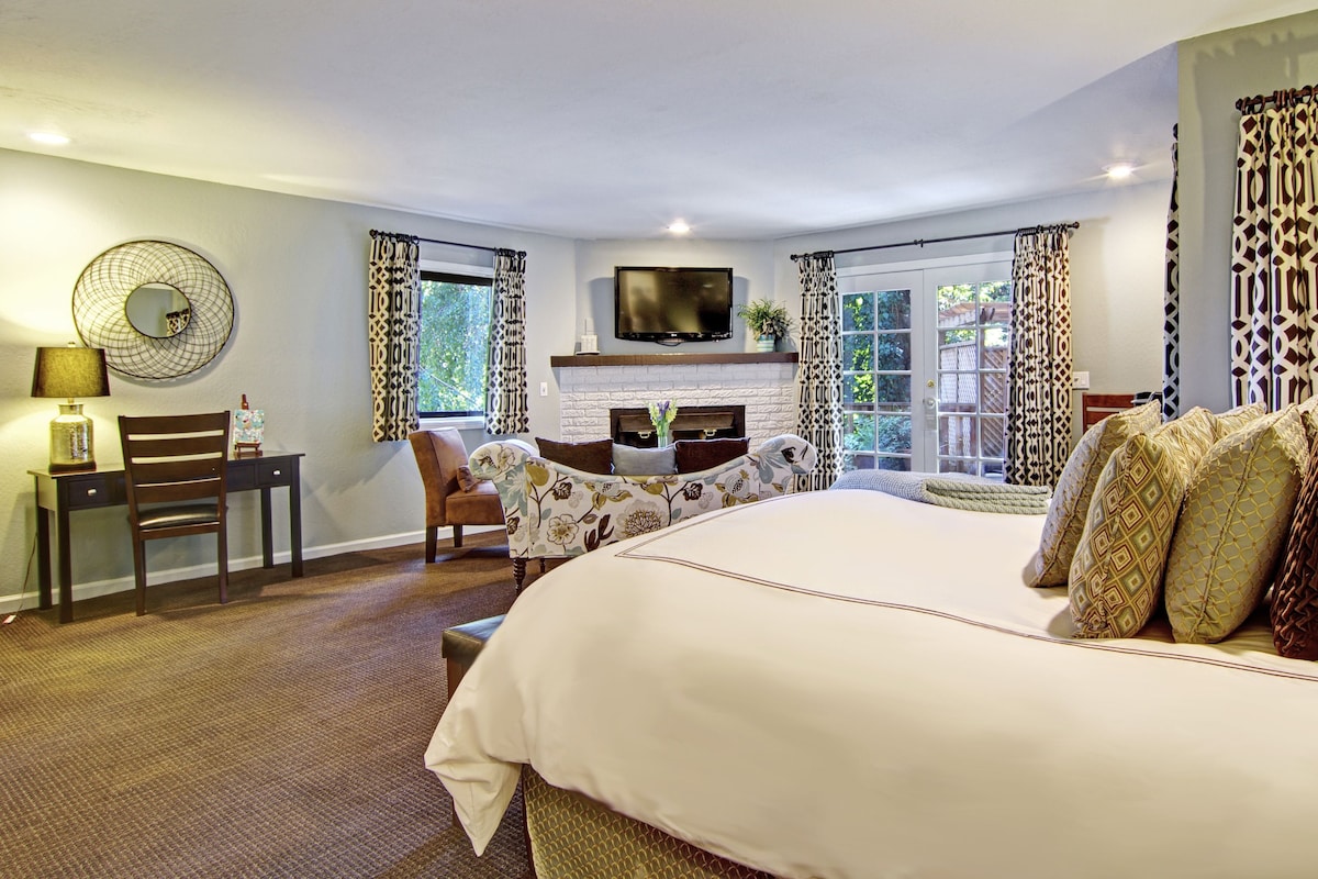 A hotel room with a large bed, a desk, a chair, a TV, and a fireplace.