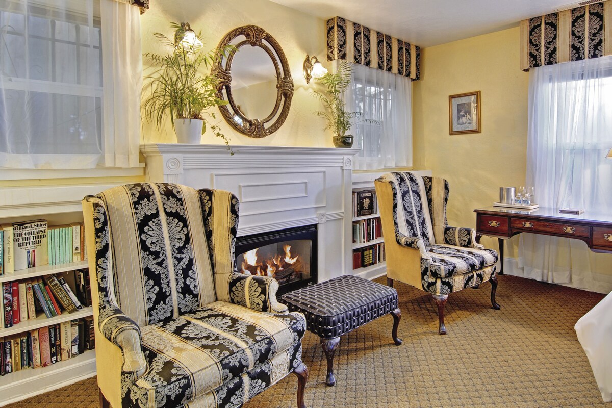 A traditional living room with a fireplace, patterned armchairs, a side table, and a bookshelf filled with books.