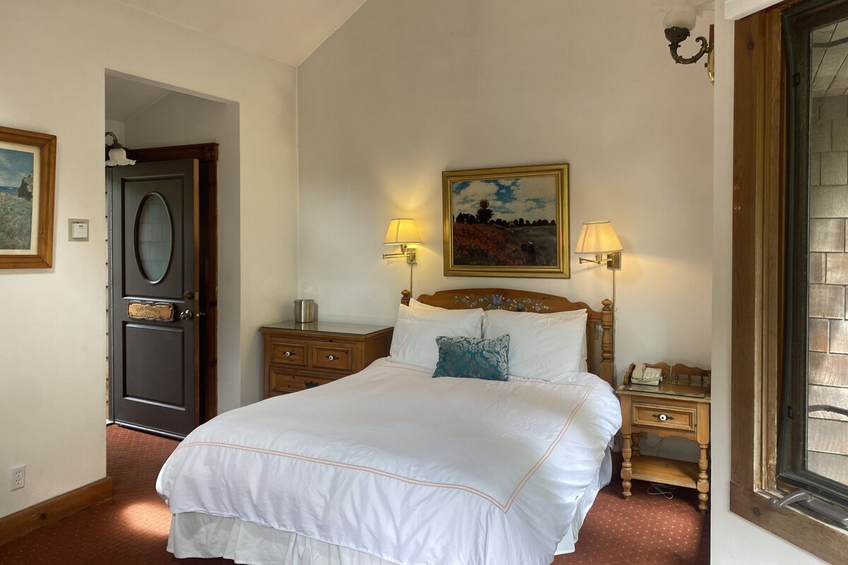 A bedroom with a large bed, two bedside lamps, a wooden nightstand, a dresser, a door, and a painting on the wall.