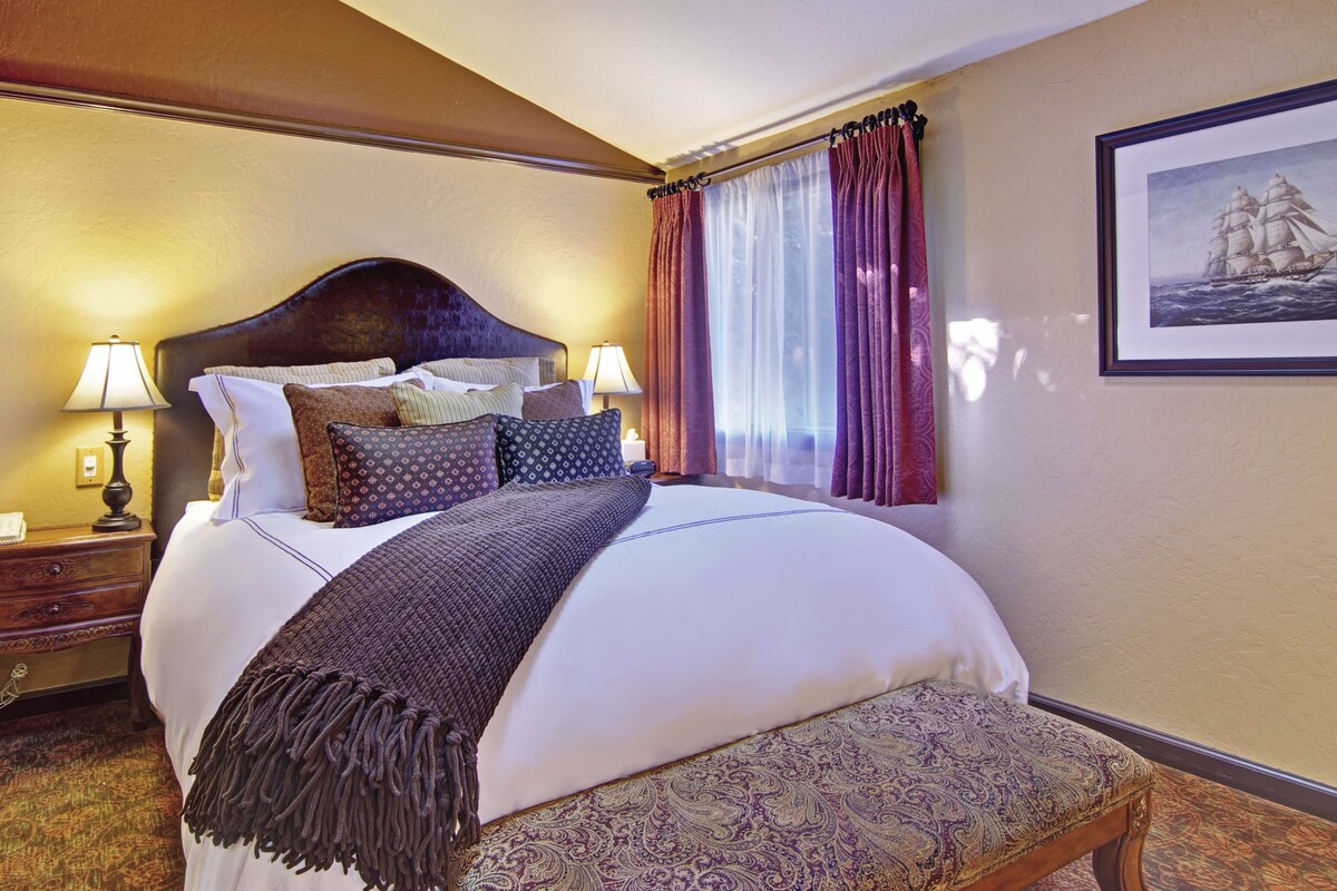 A hotel room with a large bed, two bedside tables with lamps, a bench, and a framed picture on the wall.