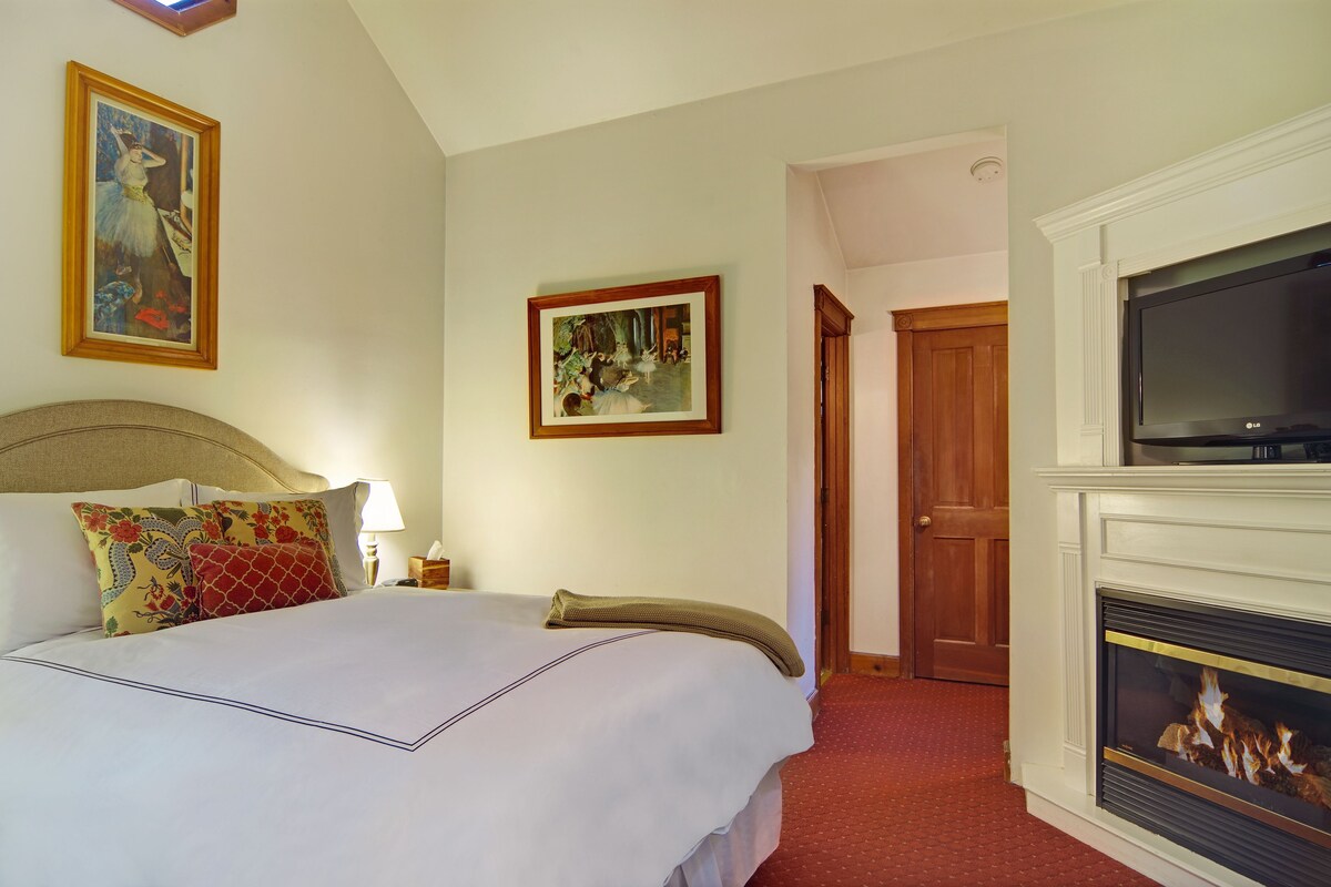 A hotel room with a bed, a TV, a fireplace, and two framed pictures on the wall.