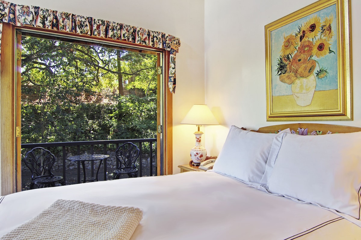 A bedroom with a large window, a bed with white linens, a nightstand with a lamp, and a painting of sunflowers on the wall.