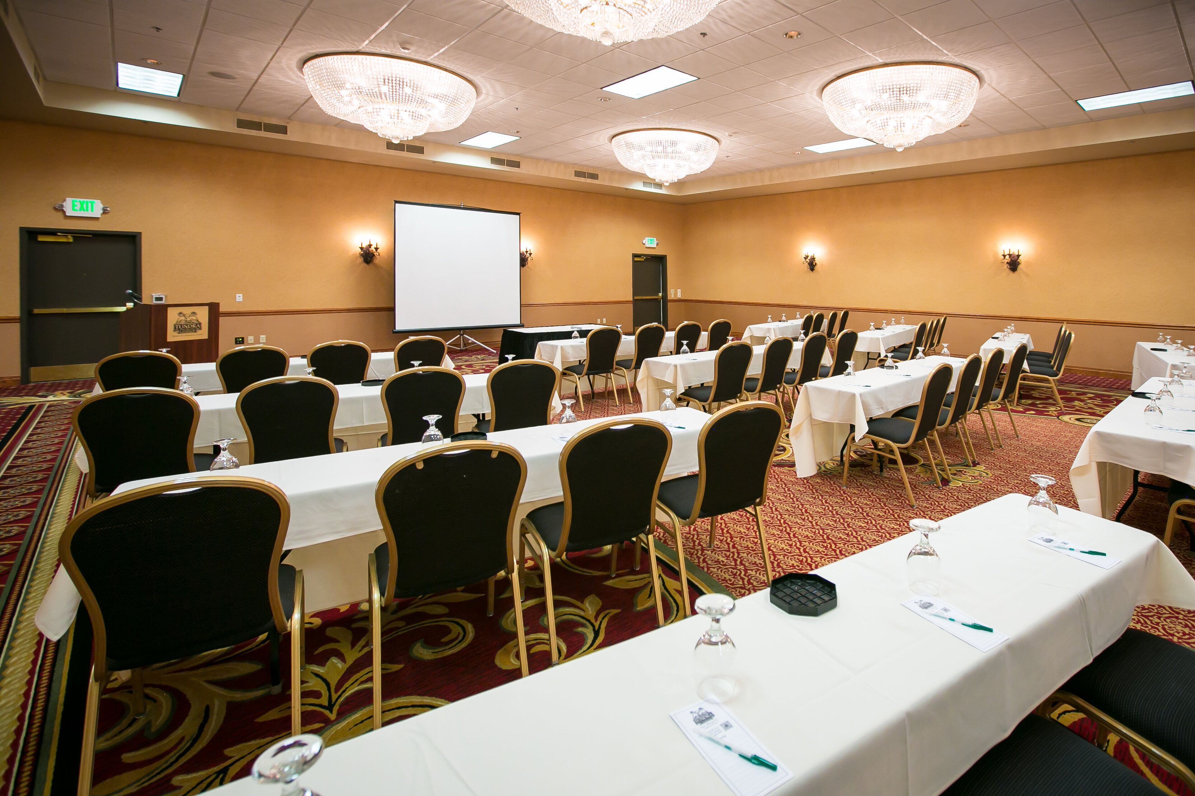 A conference room with multiple tables and chairs, a projector screen, and chandeliers.