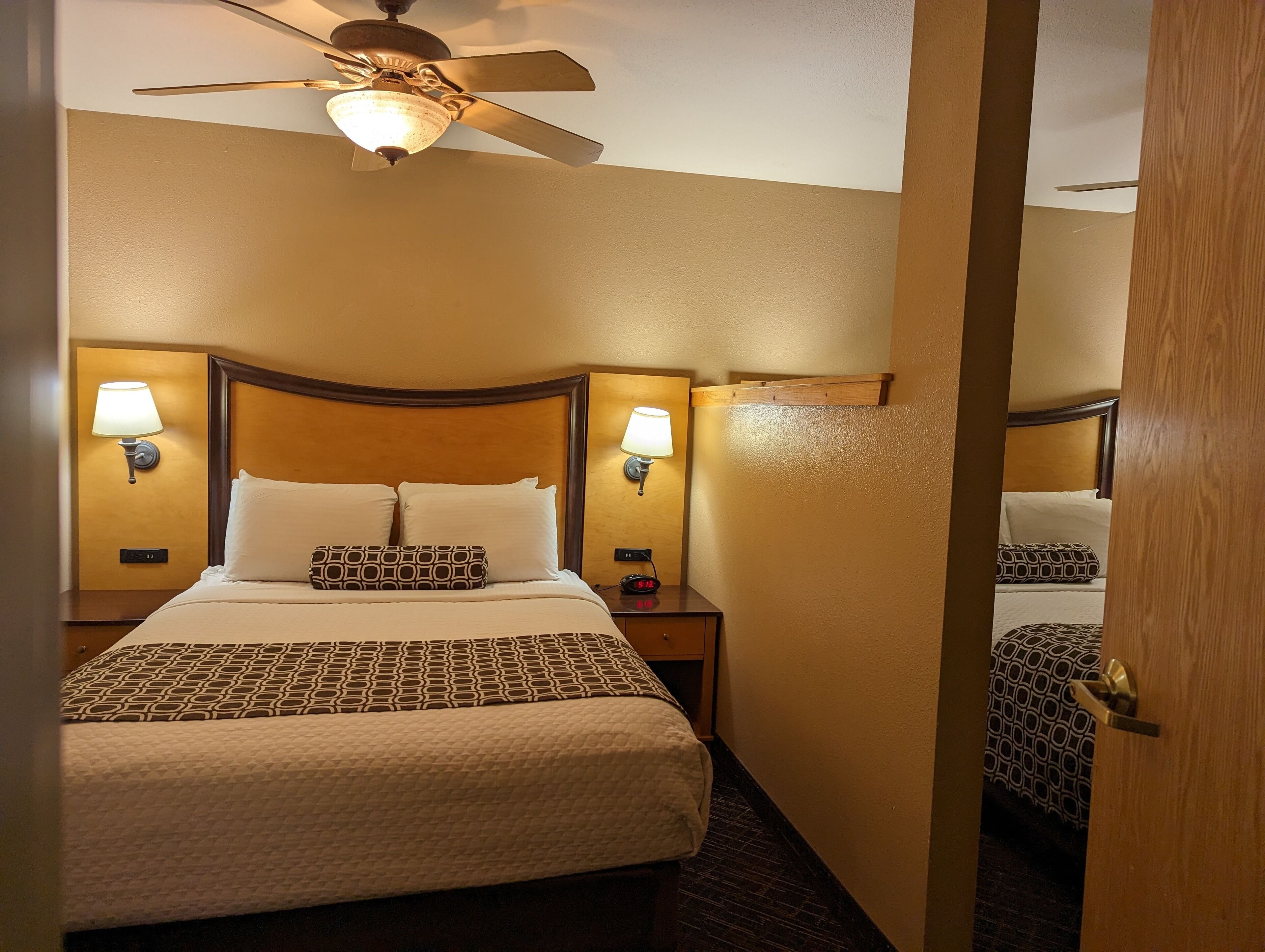 A hotel room with a bed, two bedside lamps, a ceiling fan, and a nightstand with a clock.