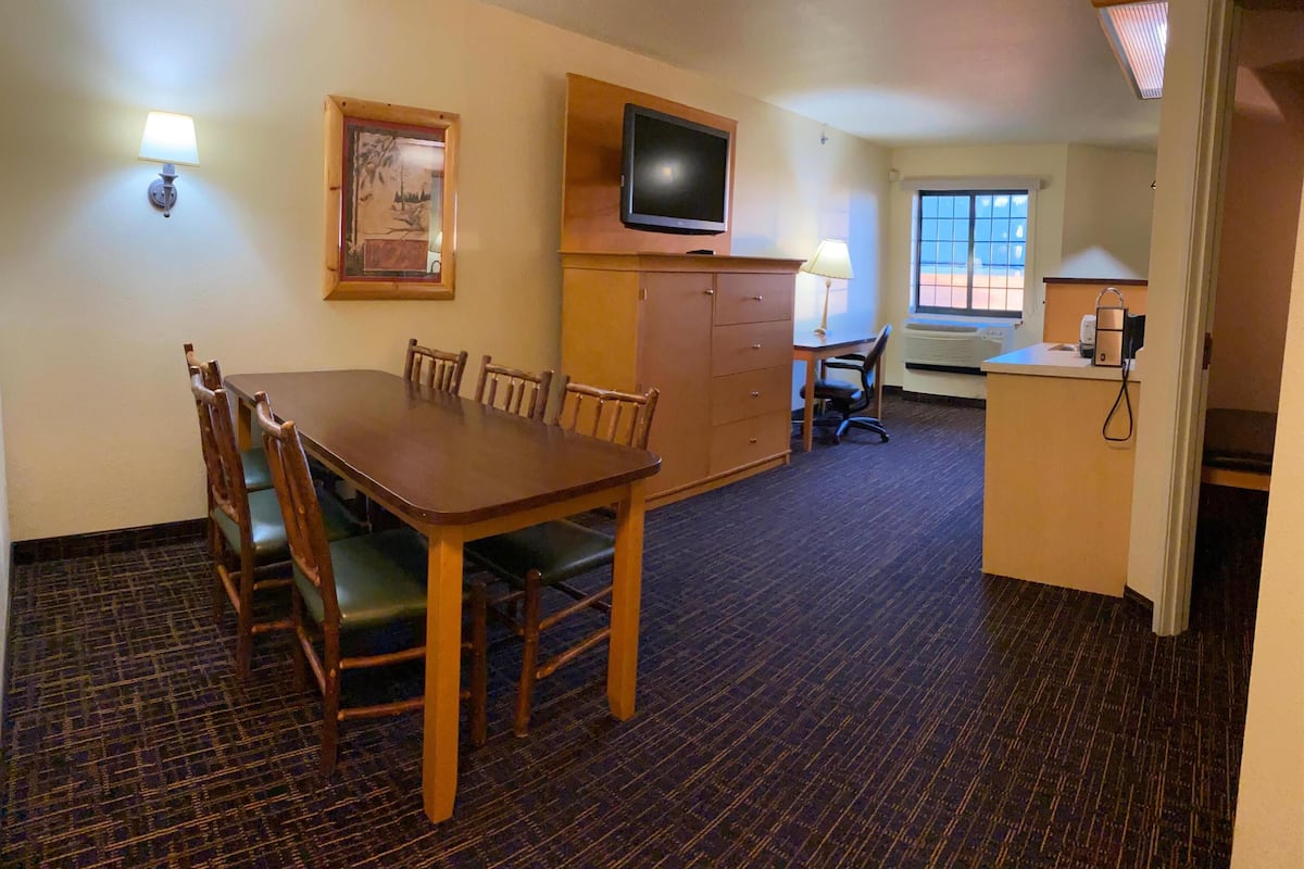 A hotel room with a dining table, chairs, a television, a desk, and a window.