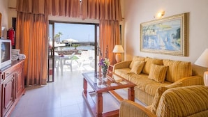 A hotel room with a sofa, a glass coffee table, a painting, and a view of the sea through a glass door. - Sands Beach Resort (Teguise)