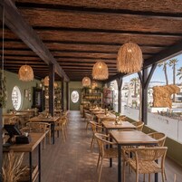 Interior de restaurante con mesas y sillas de madera, lámparas de mimbre colgantes y vistas a palmeras afuera.