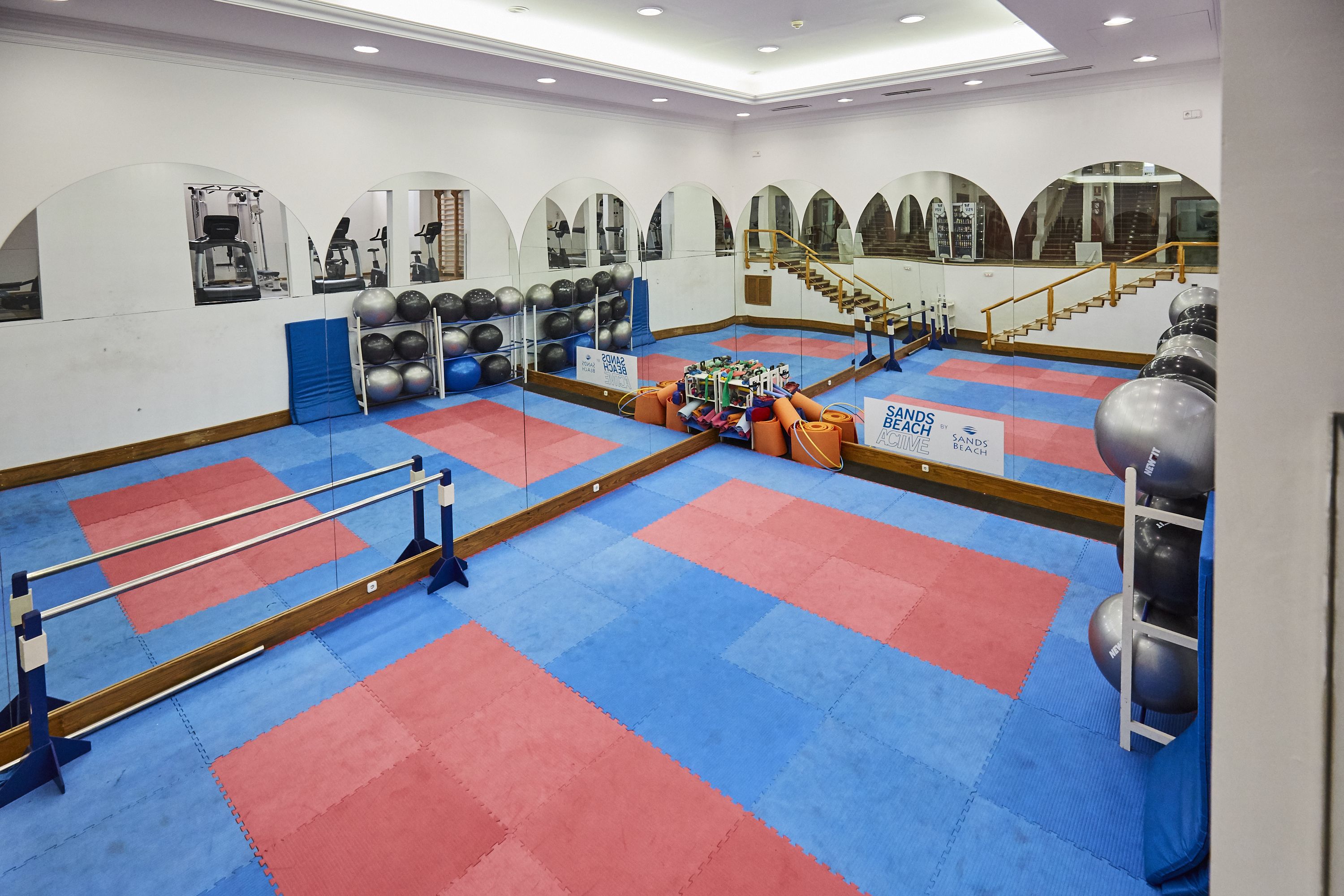Een sportschool met spiegelwanden, fitnessapparatuur en een vloer van rode en blauwe matten.